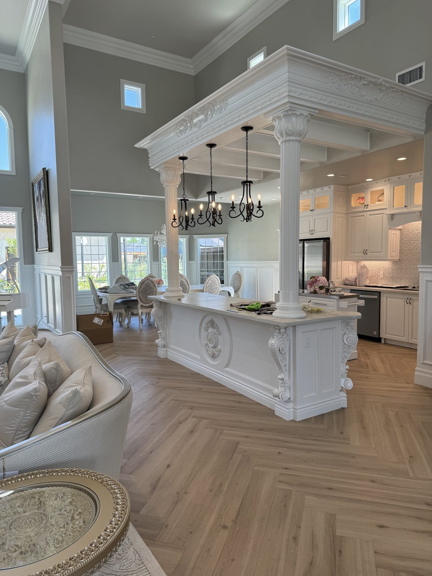 The great room of a Luxury Living home — a hand-carved island beneath Corinthian columns, wrought-iron chandeliers, and herringbone wood floors, opening to a chef's kitchen and dining area